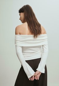 Biała bluzka z dekoltem off-the-shoulder, prążkowana, z długimi, rozszerzającymi się rękawami, w komplecie z ciemnymi spodniami. Gładka faktura i dopasowany krój.