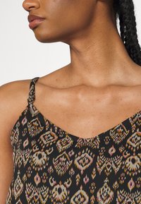 Close-up of a woman’s neck and shoulder wearing a black patterned sleeveless top with adjustable strap, hair in braids pulled back.