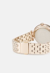 Reloj de pulsera de metal dorado que presenta un diseño de brazalete eslabonado y una caja redonda. La parte posterior es lisa y tiene una pequeña corona en el lado.