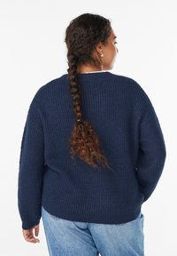 Tamsiai mėlynas megztinis su raukšlėta tekstūra, nukirptais pečiais ir laisvu siluetu. Atgalinis vaizdas rodo ilgus, laisvus rankoves ir apvalų apvadas.