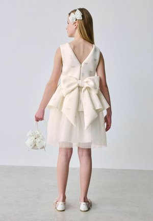 Tüdruk varrukateta elevandiluurõivas suure seljapoolse paelaga, sobiva juuksepandla, väikese käsitaskuga ja valgete kingadega, seistes ühevärvilise tausta ees.