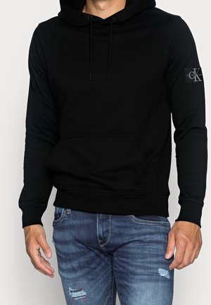 Mann trägt einen schwarzen Calvin Klein Hoodie mit Logo am Ärmel und blaue destroyed Jeans, steht mit entspannten Händen seitlich am Körper.