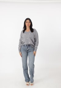 Maglione grigio con scollo a V e piccoli motivi a cuore dorati, abbinato a jeans di denim blu chiaro e sandali bianchi con tacco, su uno sfondo semplice.