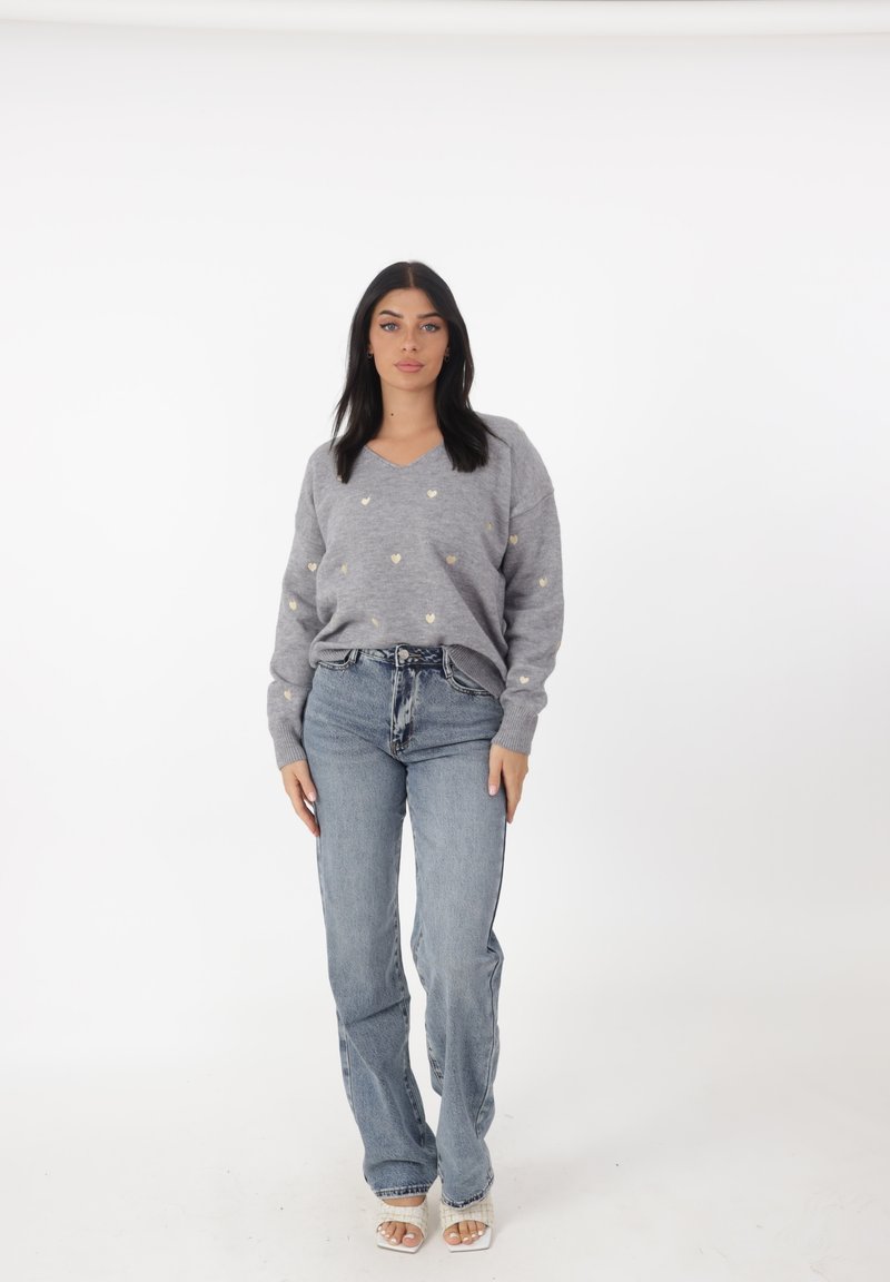 Maglione grigio con scollo a V e piccoli motivi a cuore dorati, abbinato a jeans di denim blu chiaro e sandali bianchi con tacco, su uno sfondo semplice.