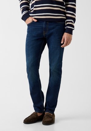 Mężczyzna w ciemnoniebieskich dżinsach, paskowanym swetrze i brązowych zamszowych mokasynach, stojący z jedną ręką w kieszeni na jednolitym tle.