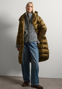 Cappotto lungo imbottito verde oliva con cappuccio, caratterizzato da una texture lucida, chiusura anteriore con zip e design imbottito, indossato sopra un cardigan di lana grigia e jeans di denim.