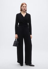 Zwarte wikkeltrui met lange mouwen, zwarte wijde broek met hoge taille en glanzende zwarte puntige schoenen. Kleine zwarte handtas met handvat.