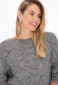 Grauer Strickpullover mit einem strukturierten Wellenmuster und offenen Gewebebereichen. Modell trägt große runde Ohrringe in Erdtönen.