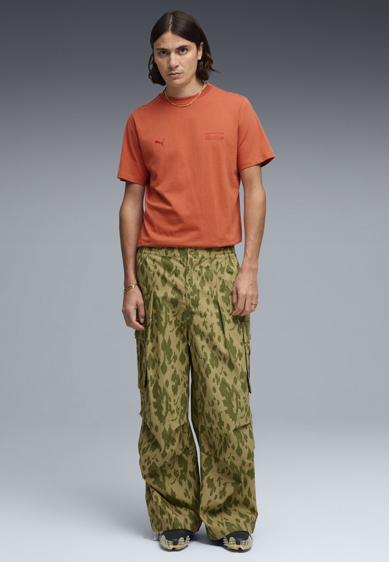 Man met schouderlang donker haar, gekleed in een oranje Puma T-shirt, olijfgroene cargobroek met patroon, en zwarte schoenen met spikes.