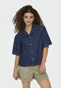 Camicia di jeans a maniche corte di colore blu scuro con bottoni dorati e cuciture a contrasto, abbinata a pantaloni corti color khaki con risvolti.