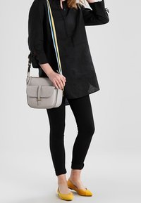 Sort tunika med krave, sorte stramme jeans, gule flade sko og en lys grå skuldertaske med en farverig rem.