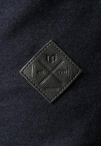 Zwarte leren logo-patch in een diamantsvorm, met stikdetails en een geperst ontwerp, op een donkere stoffen achtergrond.