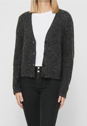 Vrouw draagt een donkergrijze geribbelde cardigan met knopen over een wit shirt, gecombineerd met een zwarte spijkerbroek met hoge taille.