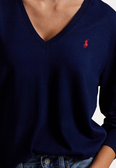 Navy blue V-neck sweater with red embroidered logo on chest, worn with blue denim jeans.