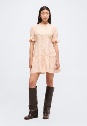 Femme portant une robe courte fleurie rose clair avec des manches à volants et un col, associée à de grandes bottes en cuir marron foncé et des boucles d’oreilles créoles dorées.