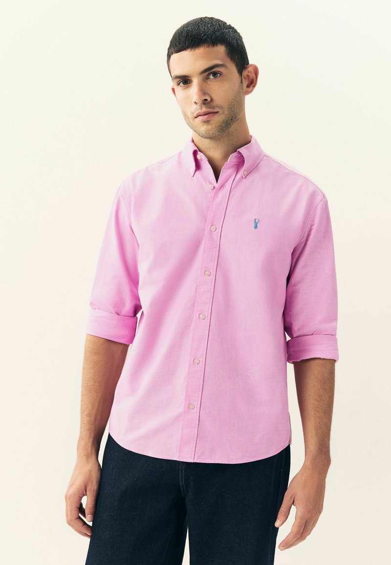 Camicia button-up rosa chiaro con maniche lunghe arrotolate fino ai gomiti, caratterizzata da un piccolo logo ricamato blu sul petto. Indossata con jeans scuri.