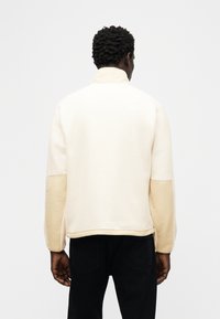 Άντρας με σκούρα σγουρά μαλλιά που φοράει μπεζ φλις μπουφάν με μπεζ επιδιορθώσεις στους αγκώνες και μαύρο παντελόνι, όρθιος με την πλάτη σε λευκό φόντο.