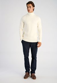 Elfenbeinfarbener Rollkragenpullover mit Rippstruktur, kombiniert mit dunkelblauen Hosen und braunen Schuhen mit hellen Schnürsenkeln. Das Model steht und hat die Hände in den Taschen.