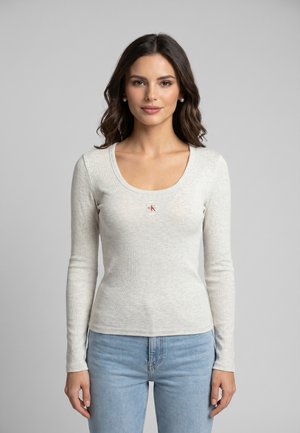 Jeune femme aux cheveux foncés portant un haut à manches longues gris clair et un jean bleu, debout devant un fond gris uni.