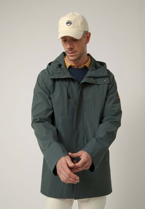 Hombre con gorra beige, chaqueta con capucha verde oscuro sobre varias capas de camisas, ajustándose las mangas contra un fondo claro y liso.