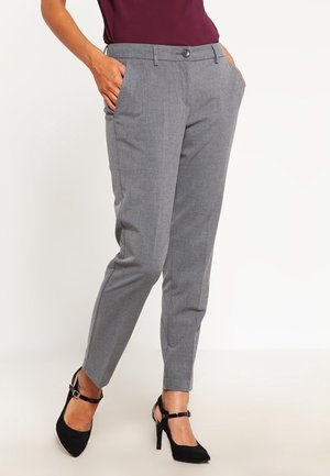 Femme portant un pantalon gris ajusté avec des poches, associé à des chaussures à talons noires et un haut bordeaux, debout devant un fond blanc.