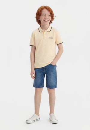 Chlapec s červenými vlasmi sa usmieva, má na sebe bledozlté polo s logom „BOSS“, rifľové kraťasy a biele tenisky, stojí s jednou rukou v kapse.