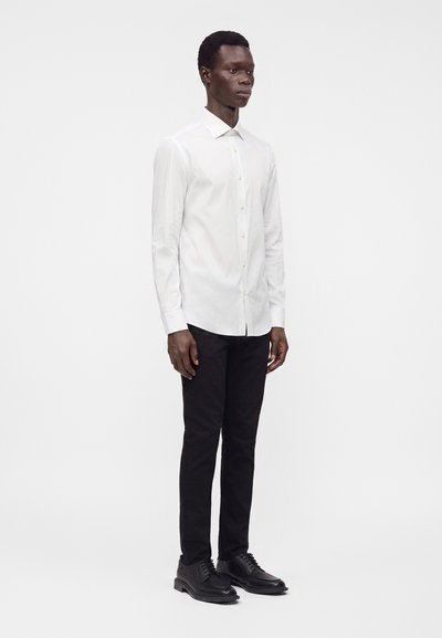 Uomo in piedi che indossa una camicia bianca a maniche lunghe con bottoni, pantaloni neri e scarpe nere stringate su sfondo bianco semplice.