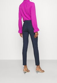 Fuchsiová blůza s vysokým límcem a volánkovými rukávy v kombinaci se zúženými námořnickými kalhotami; modelka má na sobě stříbrné sandály na vysokém podpatku.