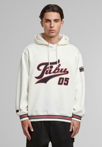 Hvit hettegenser med rød og svart "Fubu 05" grafikk, ribbet nederkant med svarte, røde og hvite striper, samt logo på ermet. Myk tekstur.
