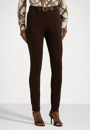 Pantalon slim marron foncé avec coutures sur le devant, associé à une chemise à motifs et des sandales à talons ouverts marron sur un mannequin debout.