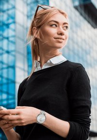 Osoba v černém svetr s bílým límečkem drží chytrý telefon, má na sobě stříbrné pletencové hodinky, s prosklenou budovou v pozadí.