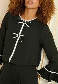 Czarna bluzka z białymi wykończeniami, z dwoma dekoracyjnymi kokardami z przodu i falbaniastymi rękawami. Gładki materiał, dopasowany krój, elegancki design.