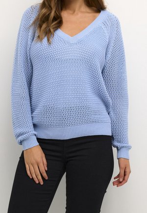 Femme portant un pull en maille bleu clair à col en V avec des manches longues et un pantalon noir, debout devant un fond uni.