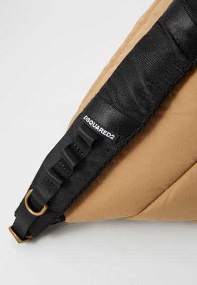 Vaatettu ruskea reppu mustalla nailonihihnalla, jossa on kultainen D-renkaalla ja merkkilabeli "DSQUARED2" hihnassa.