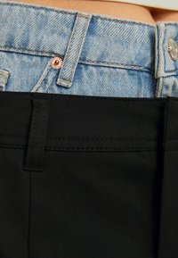 Jeans und schwarze Stoffhosen mit kontrastierenden Texturen; verfügen über einen Metallknopf und eine Naht auf dem Bund für zusätzliche Details.