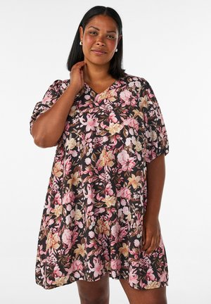 Femme aux cheveux mi-longs foncés, portant des boucles d'oreilles en perles et une robe noire à fleurs roses et beiges, se tenant devant un fond blanc.