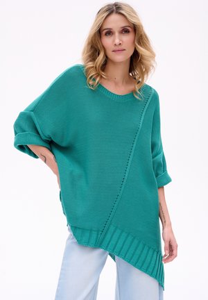 Kobieta o blond włosach, ubrana w asymetryczny turkusowy sweter dziany i jasnoniebieskie dżinsy, stoi na jednolitym jasnym tle.