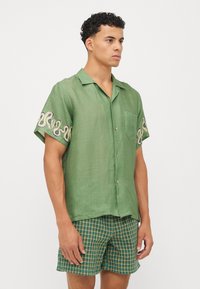 Camisa verde de manga corta con un acabado texturizado, que presenta un bordado en crema y azul marino en los mangas, combinada con pantalones cortos a cuadros.