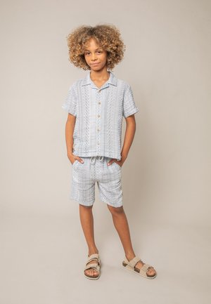 Enfant aux cheveux bouclés se tient les mains dans les poches, portant une chemise à manches courtes boutonnée gris clair à motifs, un short assorti et des sandales beiges.
