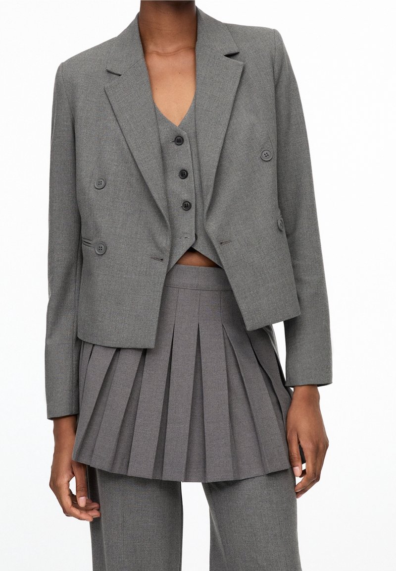Personne portant un blazer gris sur mesure, un gilet boutonné assorti, une mini-jupe plissée et un pantalon en tissu coordonné, montré du cou aux cuisses.