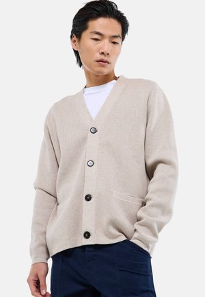 Homme portant un cardigan beige boutonné sur une chemise blanche et un pantalon bleu marine, regardant vers le bas avec les mains détendues le long du corps.