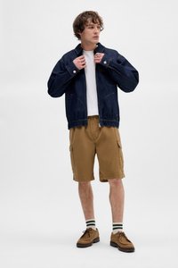 Denim jack met een rits en voorzakken, gecombineerd met khaki cargoshorts. Bruine schoenen en gestreepte sokken maken de look af. Lichte achtergrond.