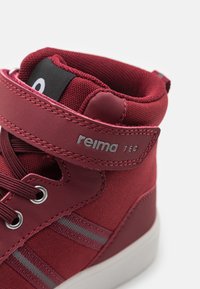 Bottines montantes rouges en toile texturée, avec une sangle velcro, des œillets métalliques et des rayures grises.