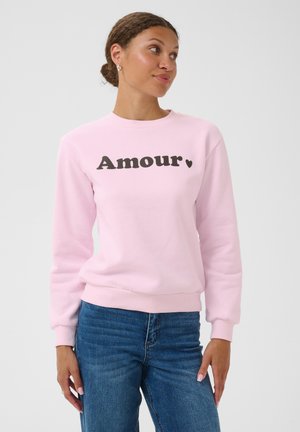Femme aux cheveux bruns attachés en arrière portant un sweat-shirt rose clair avec le mot "Amour" et un cœur, associée à un jean bleu, regardant vers la droite.