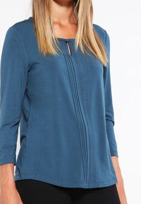 Tyrkysový dlouhý rukáv s kulatým výstřihem, detaily ve tvaru klíče, přední záhyby a mírně volný střih. Měkká, elastická tkanina.