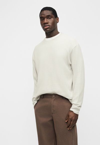 Νέος με σκούρο δέρμα που φοράει εκρού μακρυμάνικο πουκάμισο και καφέ παντελόνι, στέκεται μπροστά σε απλό ανοιχτόχρωμο φόντο.