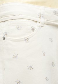 Κοντινή εικόνα σε παντελόνι από ύφασμα εκρού με floral μοτίβα σε ανοιχτό μωβ και μπεζ γύρω από τη μπροστινή τσέπη και το θηλάκι της ζώνης.