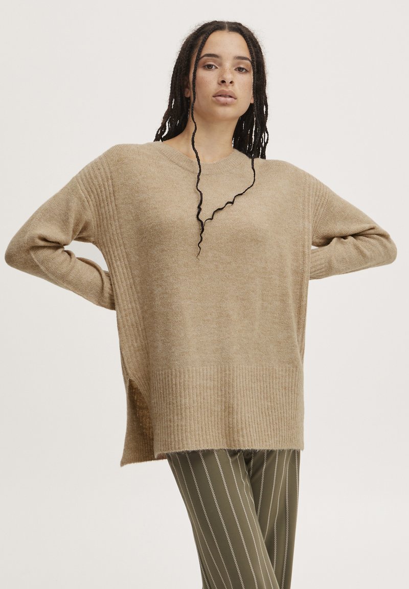 Pull en tricot beige avec une texture côtelée, coupe ample et fentes sur les côtés. Associé à un pantalon rayé vert olive.