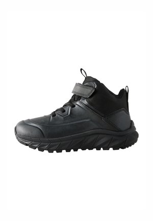 Chaussure de sport montante noire avec une semelle en caoutchouc texturée, un système de laçage, un col rembourré et une sangle Velcro autour de la cheville.