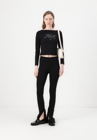 Svart langermet crop topp med "Hugo"-logo, kombinert med høy midje svarte leggings med slisse detaljer, og svarte loddrette sko.